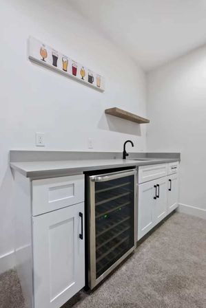 Minimalist home wet bar with white shaker cabinets, stainless glass-door wine fridge, gray countertop, matte black faucet and floating wood shelf on a white wall.