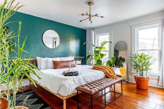 Bright modern bedroom with teal accent wall, round mirror, plush white bedding, leather tufted bench, hardwood floors, tall windows and abundant indoor plants for a cozy leafy retreat.