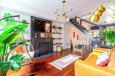 Bright modern open-concept living room with tan leather sofa, rattan and hanging chairs, large indoor plants, wall-mounted TV above a black mantel, brass pendant lights, dark hardwood floors and an open metal staircase.