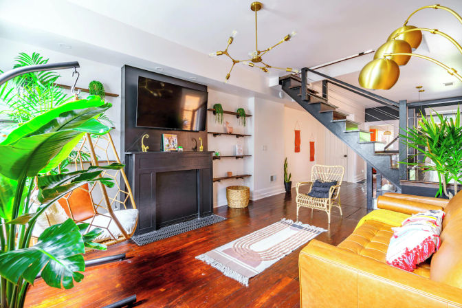 Bright modern open-concept living room with tan leather sofa, rattan and hanging chairs, large indoor plants, wall-mounted TV above a black mantel, brass pendant lights, dark hardwood floors and an open metal staircase.