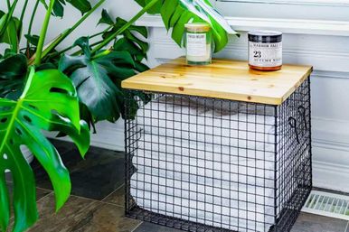 Indoor spa-style corner with large Monstera leaves beside a black wire crate side table topped with a wooden lid, stacked white towels inside and two scented candles on top.