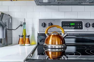 Gleaming copper tea kettle with wooden handle on a black glass electric stovetop in a modern kitchen, stainless steel range, white subway tile backsplash and countertop oil bottle and spice shakers