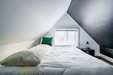 Cozy minimalist attic loft bedroom with white bedding, long fluffy bolster, green accent pillow, small window at the foot, black bedside table and lamp under sloped ceilings.