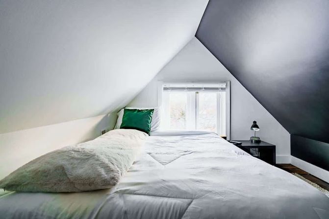 Cozy minimalist attic loft bedroom with white bedding, long fluffy bolster, green accent pillow, small window at the foot, black bedside table and lamp under sloped ceilings.