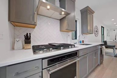 Bright modern kitchen interior with gray cabinets, white quartz countertops, stainless-steel gas cooktop and oven, angled subway-tile backsplash and overhead range hood in an open-concept home.
