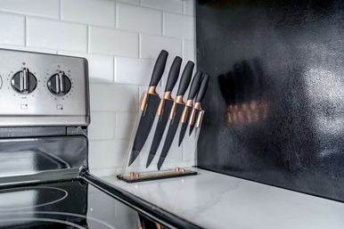 Sleek modern kitchen countertop with a clear acrylic knife block holding black-handled knives with rose-gold accents beside a stainless-steel stove and white subway-tile backsplash.