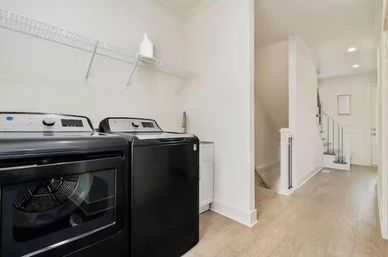 Bright residential laundry room with sleek black front-load washer and dryer under a wire wall shelf, white walls, wood-look flooring and a nearby hallway leading to a staircase.