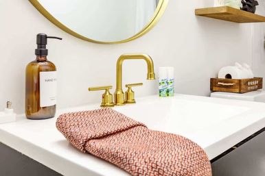 Modern home bathroom sink with brass faucet and round gold mirror, amber hand soap dispenser, coral patterned hand towel on white countertop, wooden shelf and toilet paper tray in background.