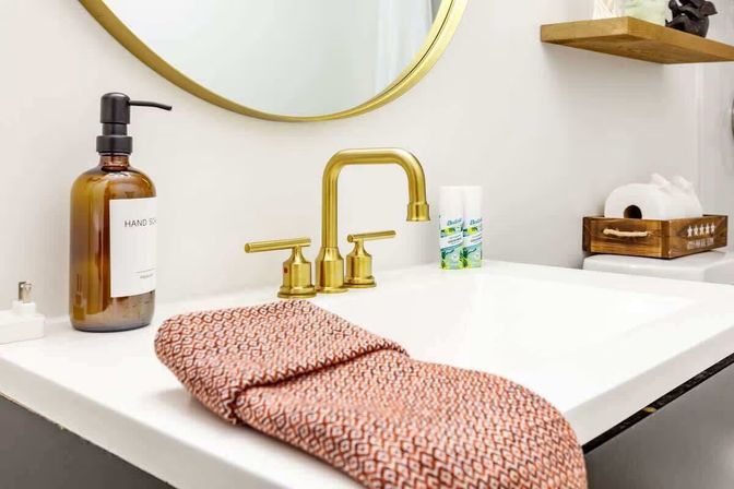 Modern home bathroom sink with brass faucet and round gold mirror, amber hand soap dispenser, coral patterned hand towel on white countertop, wooden shelf and toilet paper tray in background.