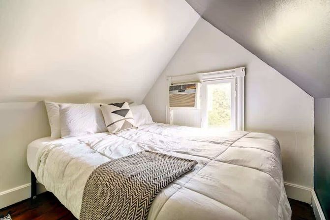 Cozy bright attic bedroom with sloped ceiling, white quilted bed, geometric throw pillow, chevron blanket and a window air conditioner