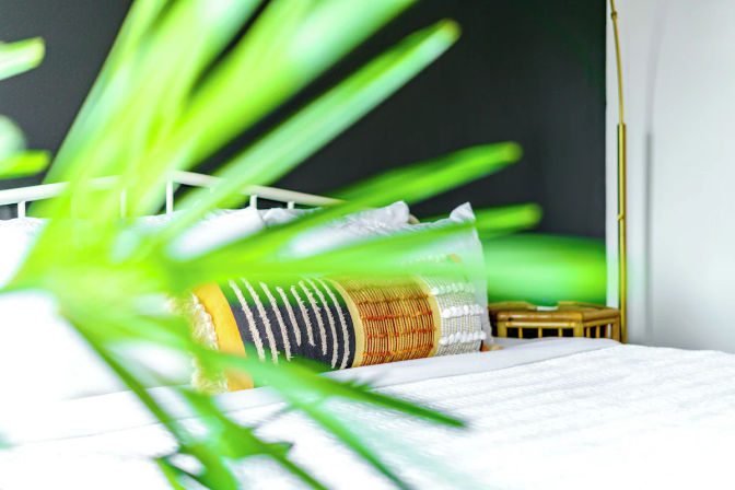 View through green palm leaves into a bright modern bedroom with white bedding, a textured black-and-gold lumbar pillow, rattan nightstand and brass floor lamp against a dark accent wall.