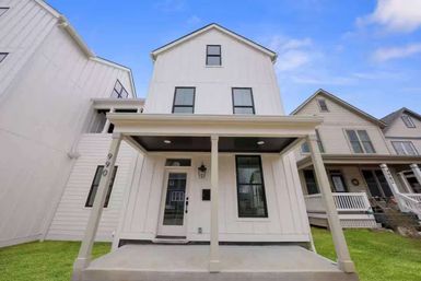 White three-story modern farmhouse with covered front porch, black-trim windows and small concrete stoop on a suburban residential street with neighboring houses and blue sky