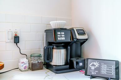 Black drip coffee maker with stainless thermal carafe on a white subway-tile kitchen counter, jar of coffee grounds, sugar packets, paper filters on top, and a tablet displaying weather (light rain, 82°F)