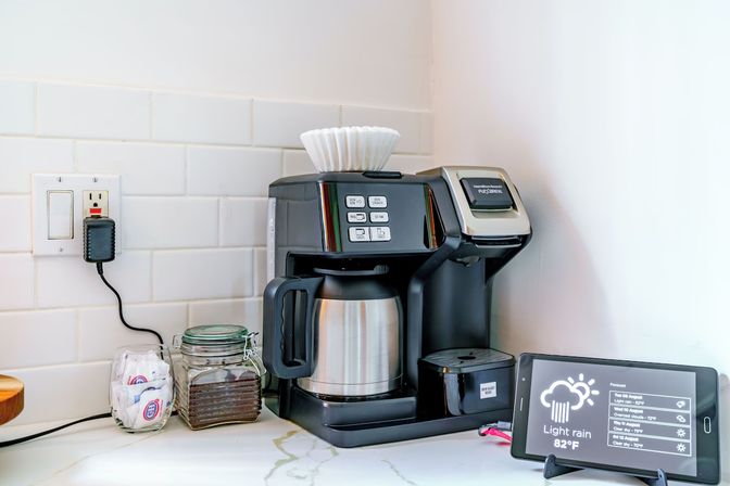 Black drip coffee maker with stainless thermal carafe on a white subway-tile kitchen counter, jar of coffee grounds, sugar packets, paper filters on top, and a tablet displaying weather (light rain, 82°F)