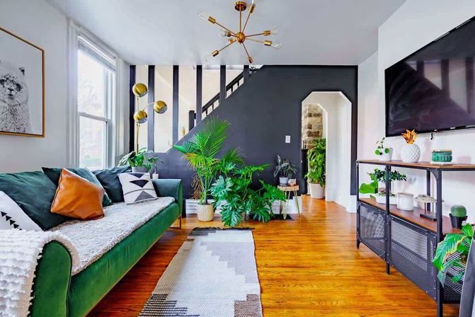 Sunlit modern living room with a green velvet sofa and leather accent pillow, black accent wall by the stairs, warm hardwood floors, abundant indoor plants, gold mid-century chandelier, patterned rug, and wall-mounted TV.