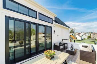 Sunlit rooftop deck of a modern white house with black-framed sliding doors and clerestory windows, wooden picnic table with potted flowers, wicker lounge seating and coffee tables overlooking a suburban neighborhood under a blue sky.
