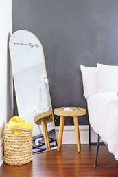 Cozy modern bedroom corner with a gold-framed full-length mirror leaning against a gray accent wall, round wicker stool and woven basket with a yellow blanket on warm hardwood floors beside a white bed