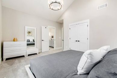Bright modern master bedroom with gray linens, white dresser and full-length mirror, vaulted ceiling light fixture, double closet doors and open doorway to an ensuite bathroom