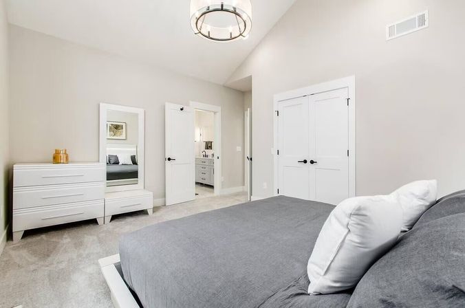Bright modern master bedroom with gray linens, white dresser and full-length mirror, vaulted ceiling light fixture, double closet doors and open doorway to an ensuite bathroom