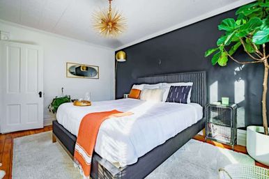 Sunlit, cozy modern bedroom with a charcoal accent wall, gray upholstered bed dressed in white linens and an orange throw, gold sputnik chandelier, fiddle‑leaf fig plant, wire bedside table and warm hardwood floors.