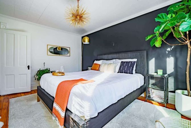 Sunlit, cozy modern bedroom with a charcoal accent wall, gray upholstered bed dressed in white linens and an orange throw, gold sputnik chandelier, fiddle‑leaf fig plant, wire bedside table and warm hardwood floors.