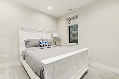 Bright minimal bedroom interior with a white paneled bed frame, gray bedding, geometric blue accent pillow, bedside lamp and a window with blinds.