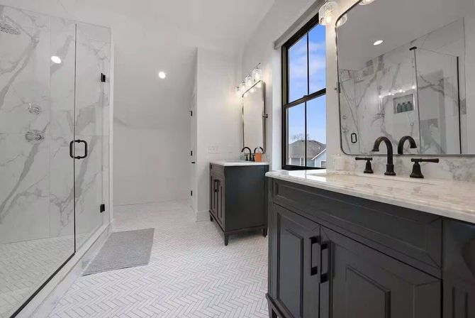 Modern bright bathroom with dark wood double vanities, marble countertops and black faucets, large frameless glass walk-in shower with marble tile, white herringbone floor tile and a window overlooking suburban rooftops.