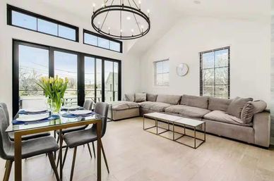 Bright, airy open-plan living and dining area with light wood floors, gray sectional sofa, glass dining table with yellow tulips, large black-framed sliding doors and round chandelier.