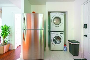 Bright, compact modern laundry nook with stacked white washer and dryer in a recessed alcove beside a two-door stainless steel refrigerator, marble-look tile floor, trash bin, detergent bottle, and potted plant by a hardwood-floor hallway.