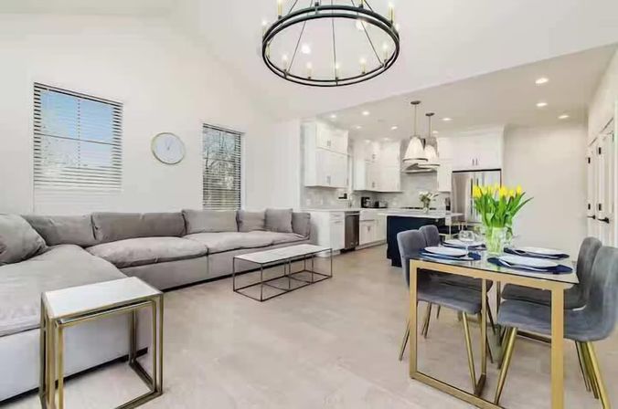Bright modern open-concept living room and kitchen with a large gray sectional, minimalist coffee table, gold-accent dining table set with yellow tulips, white cabinets, kitchen island, and round chandelier