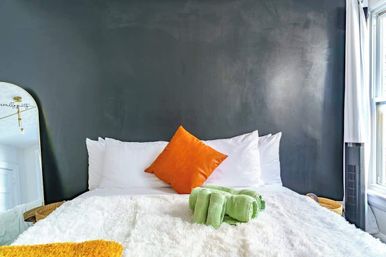 Cozy modern bedroom with dark gray accent wall, white fluffy bedding, bright orange throw pillow, green rolled towels, full-length mirror and sunlit window