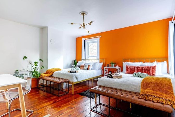 Sunny, modern bedroom with two beds against a bold orange accent wall, warm hardwood floors, tufted leather benches, white bedding with colorful throw pillows and cozy throws, potted plants and a window letting in natural light.