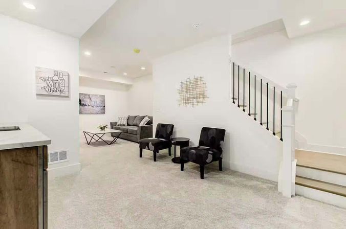 Finished lower-level living room with light carpet, gray sofa and geometric coffee table, two patterned black accent chairs and round side table by a white staircase with black balusters, modern wall art and recessed lighting.