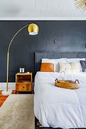 Modern bedroom interior with matte black accent wall, gold arched floor lamp, wooden nightstand, white duvet bed with leather accent pillow and wooden tray — cozy minimalist bedroom decor.