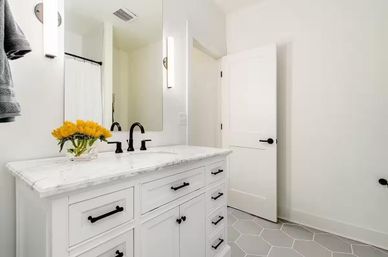 Bright modern white bathroom with marble-top vanity, matte black faucet and hardware, gray hexagon floor tiles, sunflower bouquet in a glass vase, wall mirror and open white door.