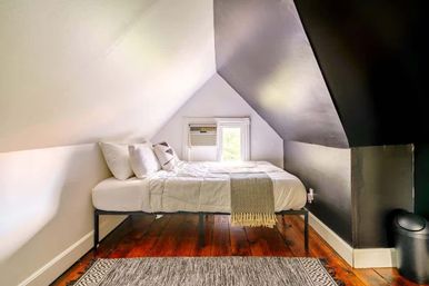 Cozy attic loft bedroom with sloped ceilings, white-bedded platform bed, window air conditioner, dark accent wall, warm hardwood floors and patterned rug.