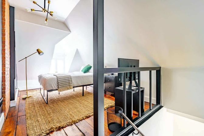 Sunlit attic loft bedroom with exposed brick, hardwood floors and woven jute rug, metal-framed bed with white linens and green pillow, brass floor lamp, modern chandelier and black railing by the staircase