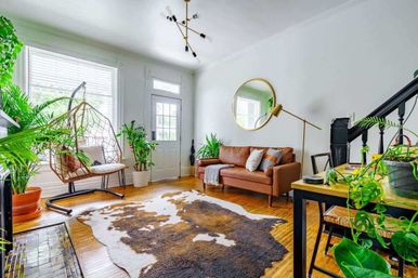 Bright sunlit boho living room with a tan leather sofa, hanging wicker swing chair, abundant potted plants, round wall mirror, and cowhide rug on hardwood floors.