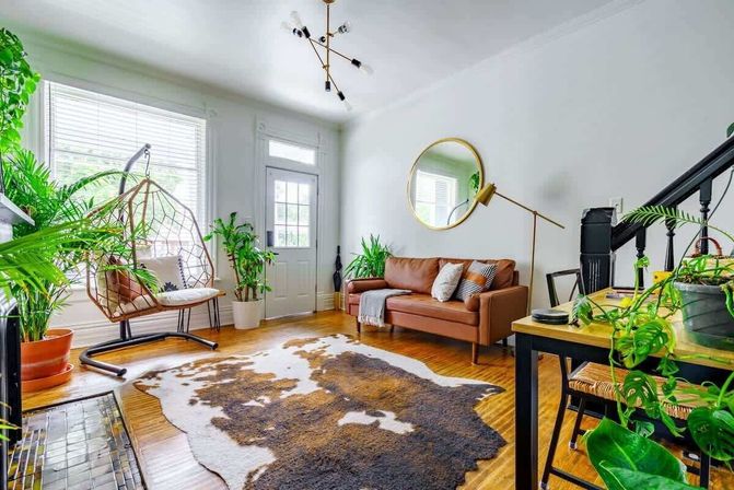 Bright sunlit boho living room with a tan leather sofa, hanging wicker swing chair, abundant potted plants, round wall mirror, and cowhide rug on hardwood floors.