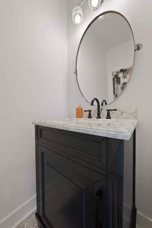 Compact powder room vanity with black cabinet, marble countertop, matte black faucet, round wall mirror and amber soap dispenser beneath glass sconce lights