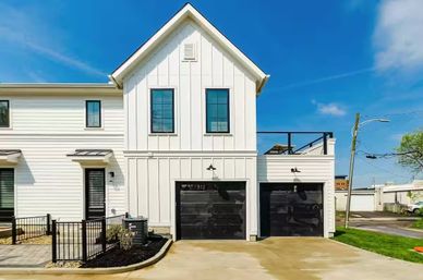 Modern white two-story farmhouse-style duplex with board-and-batten siding, dual black garage doors, rooftop terrace, small fenced patio, paved driveway and bright blue sky in a suburban residential area.