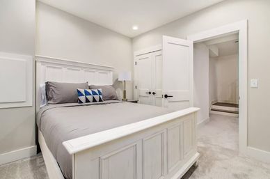 Modern farmhouse-style bedroom with white paneled wood bed, gray bedding, blue geometric accent pillow, bedside lamp, neutral walls, recessed light and open doorway to a carpeted hallway.