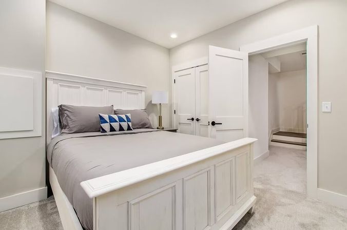 Modern farmhouse-style bedroom with white paneled wood bed, gray bedding, blue geometric accent pillow, bedside lamp, neutral walls, recessed light and open doorway to a carpeted hallway.