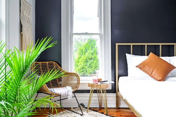 Modern boho bedroom interior with a black accent wall, gold metal bed frame, white linens and tan pillow, rattan chair, indoor palm plant, wooden side table and sunlit window.