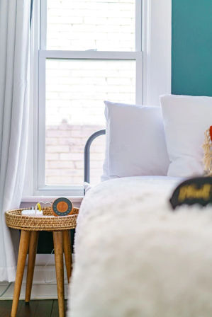 Cozy sunlit bedroom corner with white bedding and pillows, teal accent wall, wicker round side table with wooden legs holding a small orange portable speaker and charger by a bright window.