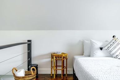 Bright minimalist attic bedroom with white quilted bed and patterned pillows, small bamboo bedside table, wicker towel basket, metal railing and dark hardwood floor