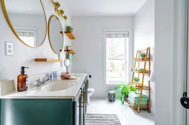 Cheerful sunlit modern home bathroom with green double-sink vanity, gold round mirrors, wooden floating shelves and a bamboo ladder shelf stacked with towels and potted plants by a bright window