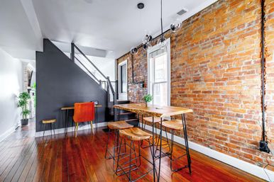 Loft-style urban dining and workspace with exposed brick wall, hardwood floors, live-edge wood table and metal stools, orange accent chair and glass globe pendant lights.