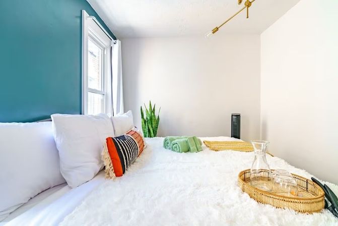 Sunlit modern bedroom with teal accent wall, fluffy white bedding, colorful striped throw pillow, snake plant by the window, green rolled towels and a wicker tray with glass carafe on the bed.