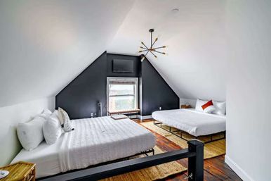 Cozy attic loft bedroom with two queen beds in white linens, dark triangular accent wall, sputnik chandelier, hardwood floors, woven rugs and a window bench.